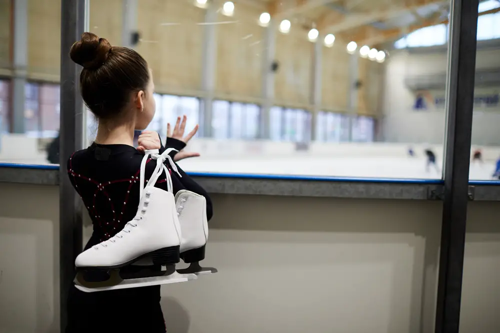 Back view portrait of little girl holding figure skates standing by ice rink and watching training